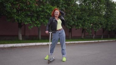 Happy confident beautiful retro woman with kick scooter standing on street road laughing looking at camera. Wide shot portrait beautiful Caucasian lady posing in 1980s 1990s in town in slow motion.