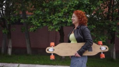 Tracking shot confident beautiful redhead retro woman walking in slow motion with skateboard smiling. Portrait of positive attractive Caucasian lady strolling on town street outdoors enjoying leisure.