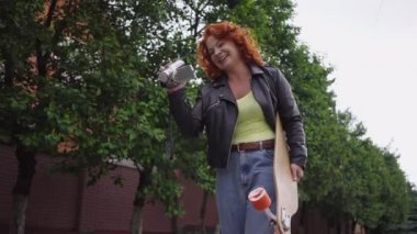 Confident redhead Caucasian retro woman walking with skateboard filming on vintage camera smiling. Tracking shot portrait beautiful fit lady strolling in town outdoors enjoying hobby. Slow motion.