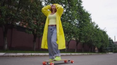 Beautiful relaxed retro woman putting on yellow rain coat standing with skateboard outdoors on street road. Wide shot portrait of cheerful Caucasian confident lady enjoying leisure on spring day.
