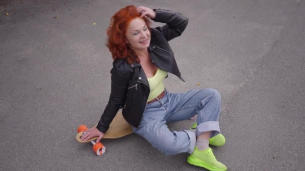 High angle view portrait attractive retro woman touching hair looking at camera sitting on skateboard on road. Wide shot happy confident Caucasian lady posing in 1980s 1990s outdoors in slow motion. — Stockvideo