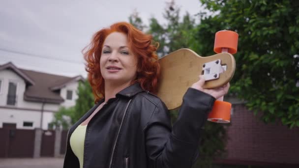 Portrait of confident gorgeous redhead retro woman posing outdoors with skateboard on shoulders. Beautiful Caucasian lady standing on town street looking at camera and looking away. — Stok video