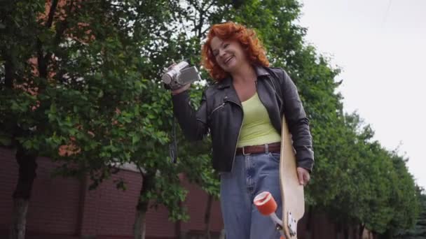 Confident redhead Caucasian retro woman walking with skateboard filming on vintage camera smiling. Tracking shot portrait beautiful fit lady strolling in town outdoors enjoying hobby. Slow motion. — Vídeo de Stock
