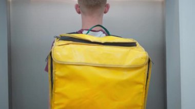 Back view young male courier with yellow thermal bag entering elevator in house or business center. Caucasian man delivering takeaway food walking in lift indoors. Delivery service concept.