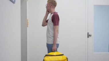 Side view portrait of young Caucasian man talking on phone waiting for lift with yellow food delivery bag. Courier delivering takeaway order standing indoors hanging up pressing elevator button.