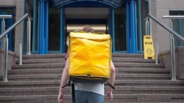Confident young courier walking upstairs entering business center in urban city. Back view wide shot Caucasian man with yellow insulated thermal food delivery bag outdoors delivering lunch order.