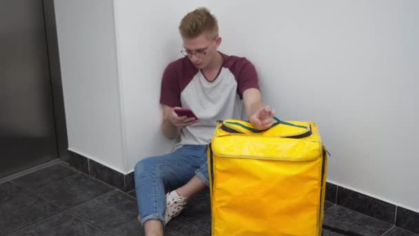 Portrait of young handsome Caucasian man sitting on floor with yellow thermal backpack surfing Internet on smartphone. Male courier waiting for client in business center or residential building. — Vídeo de Stock