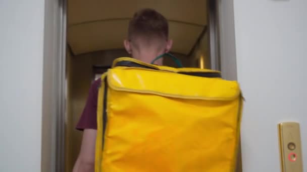 Tracking shot young man entering lift with yellow insulated bag turning pressing button. Live camera follows Caucasian male courier with food delivery backpack walking in elevator. — Video Stock