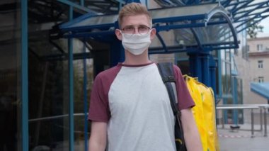 Rack focus from male courier in eyeglasses and coronavirus face mask to stretched package with takeaway food. Portrait of Caucasian young man delivering order on pandemic in urban city outdoors posing