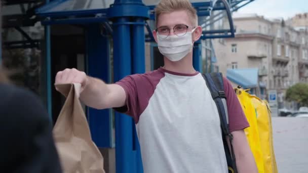 Caucasian man in coronavirus face mask and eyeglasses passing food delivery parcel to woman outdoors at business center. Positive courier talking to client crossing hands smiling. Urban delivery. — Vídeo de stock