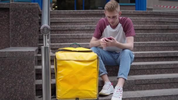 Wide shot portrait of concentrated Caucasian courier sitting on urban stairs with smartphone using app. Live camera zoom in to yellow insulated thermal backpack with takeaway food. — Wideo stockowe