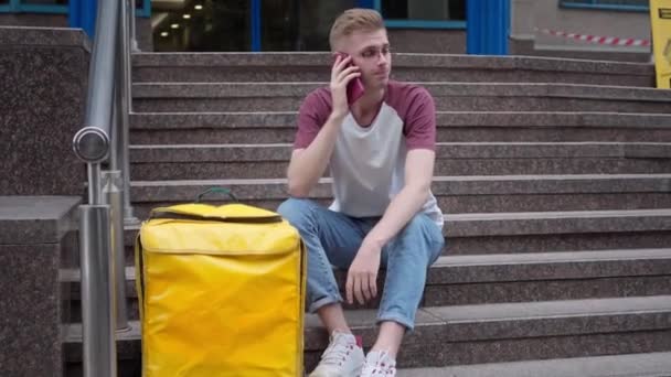 Young Caucasian courier in eyeglasses sitting on stairs outdoors talking on phone. Portrait of confident handsome man with yellow thermal backpack discussing takeaway food delivery with client waiting — Video Stock