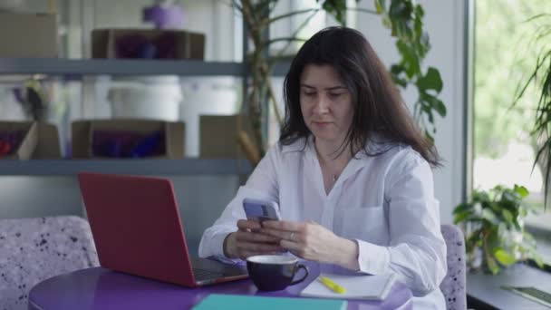 Vážná kavkazská dospělá žena sedící v kavárně s notebookem a přemýšlející o zasílání zpráv online na smartphonu. Portrét koncentrované freelancer surfing Internet v kavárně uvnitř. Zpomalený pohyb. — Stock video