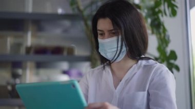 Coronavirus 'ta odaklanmış beyaz kadın yüz maskesi takıyor. Tablet üzerinde internette sörf yapıyor. Kapalı kafede oturuyor. Covid-19 'daki kafede internetten mesajlaşan konsantre akıllı yöneticinin portresi..