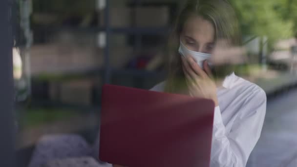 Mladá žena v koronavirus tvář maska kašlání surfování na internetu na notebooku sedí v kavárně uvnitř. Portrét nemocné kavkazské na volné noze zasílání zpráv online v kavárně s příznaky onemocnění. — Stock video