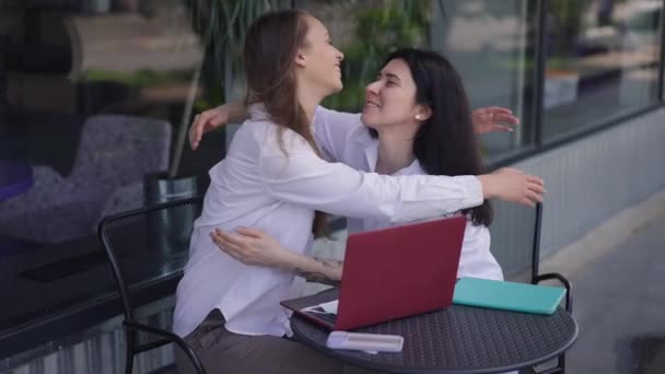 Jóvenes mujeres felices abrazándose en cámara lenta sentado a la mesa al aire libre en la terraza de verano cafetería. Amigas caucásicas sonrientes positivas abrazando la reunión para el almuerzo en la ciudad urbana. Estilo de vida y amistad — Vídeos de Stock