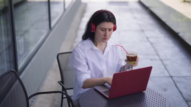 Koncentrerad kvinna i hörlurar surfar Internet på laptop sitter vid bordet med uppfriskande frukt cocktail utomhus. Porträtt av upptagen vit dam meddelande online på lunch i café sommar terrass. — Stockvideo