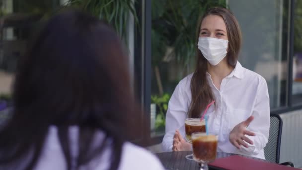 Young woman in Covid face mask talking with friend sitting at table in cafe summer terrace looking around smiling. Positive happy relaxed Caucasian millennial lady gossiping outdoors in urban city. — Stock Video