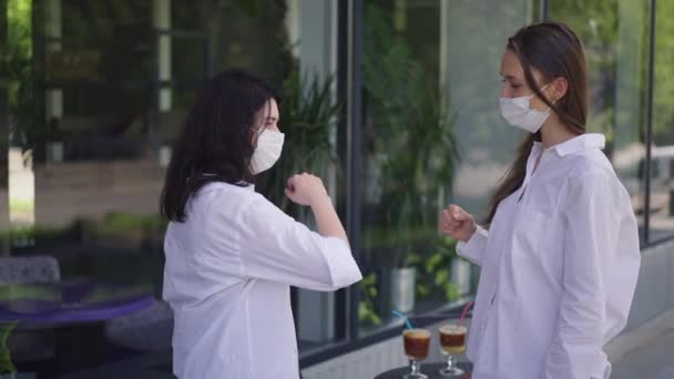 Dua wanita dengan masker coronavirus menyentuh siku dalam gerakan lambat berbicara tentang pertemuan di luar ruangan di kafe trotoar. Pandangan dari sudut pandang orang Kaukasia yang positif mengobrol di luar ruangan di jalan kota.. — Stok Video