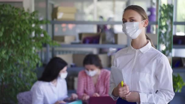 Ritratto di giovane cameriera fiduciosa positiva in maschera facciale Covid in posa in caffè con clienti felici sullo sfondo. Bella donna caucasica che guarda la macchina fotografica sorridente. Riapertura attività ristorazione. — Video Stock