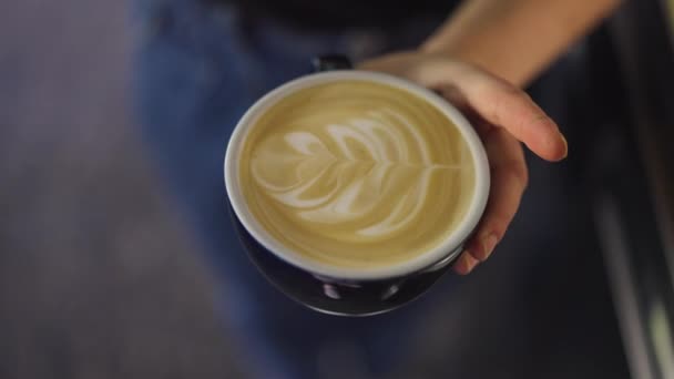 Zbliżenie cappuccino filiżanka kawy z postaci liści rysowane z widokiem na mleko góry. Smaczny poranny napój w kobiecej, białej ręce. Koncepcja sztuki latte. Zwolniony ruch. — Wideo stockowe