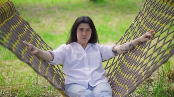 Portret van een gelukkige vrouw die in een hangmat zit te zwaaien en naar de camera kijkt. Blanke zorgeloze duizendjarige dame genieten van de lente zomer vrije tijd in het park op vakantie buiten. Langzame beweging. — Stockvideo