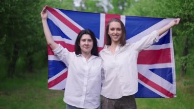 İngiliz bayrağını ağır çekimde saran mutlu kadınlara zum yap. Yazın parkta poz veren ve gülümseyen gururlu beyaz arkadaşlar..