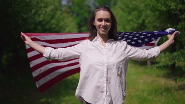 Портрет гордой худой молодой женщины, обернутой флагом США, смотрящей на улыбающуюся камеру. Живая камера увеличит изображение красивой дамы тысячелетия, позирующей в замедленном движении на открытом воздухе в весеннем летнем парке. — стоковое видео
