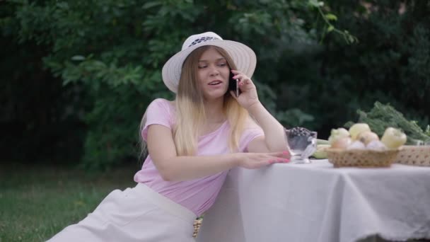 Medium shot Caucasian young woman talking on phone laughing sitting on backyard with ripe organic berries and vegetables on table. Positive beautiful lady in spring summer garden. Lifestyle concept. — Stock Video