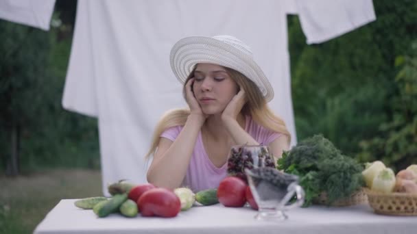 Közepes felvételű portré fiatal, szomorú nőről, aki a kertben ül az asztal előtt zöldségekkel gondolkodva. Fáradt kimerült kaukázusi női mezőgazdasági termelő szabadban, ökológiai termeléssel. — Stock videók