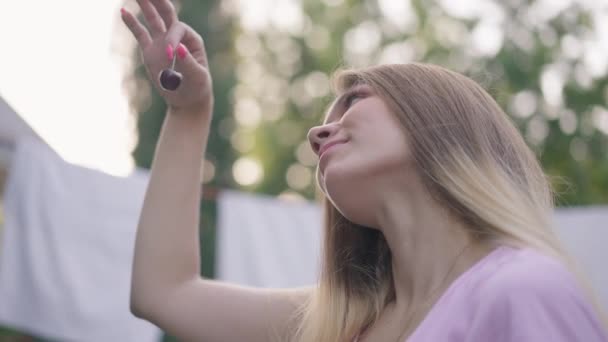 Side view of slim charming woman admiring ripe cherry sitting outdoors in garden. Portrait of millennial Caucasian lady enjoying sunny summer day outdoors on backyard with berry. Slow motion. — Stock Video