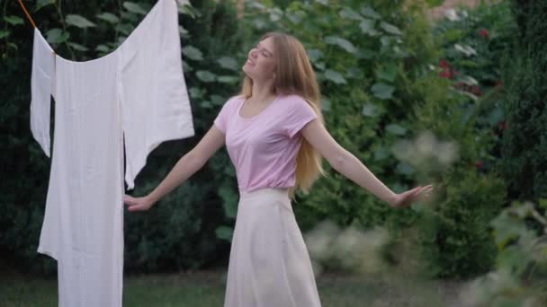 Happy young woman spinning on backyard with clean white laundry hanging. Portrait of positive Caucasian slim beautiful rural lady having fun outdoors on spring summer day. Housekeeping lifestyle. — Stock Video