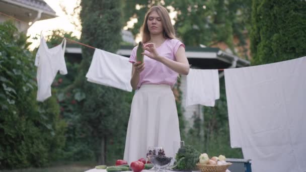 Porträtt av ung kvinna som skalar majs i slow motion stående på bakgården med ren tvätt hängande på rep och färska grönsaker liggande på bordet. Självsäker vit hemmafru på sommardagen utomhus. — Stockvideo