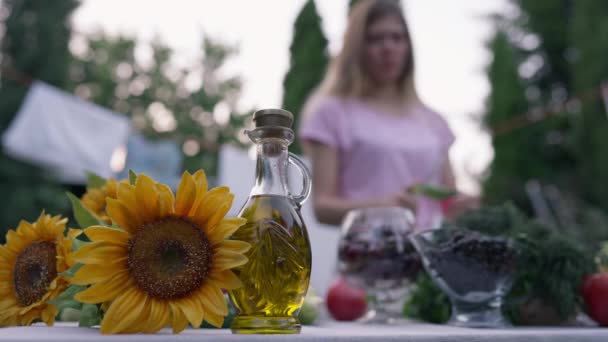 Close-up plantaardige olie en zonnebloem op tafel met wazig jonge slanke vrouw koken salade op de achtergrond. Biologische bakolie en blanke dame die komkommer snijdt in slow motion zittend op de achtertuin. — Stockvideo