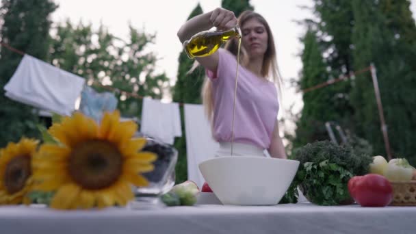 Matlagning olja häller i slow motion i vit skål utomhus på sommaren vårdagen. Suddig ung vit kvinna dressing sallad med vegetabilisk olja på bakgården. Matlagning och livsstil. — Stockvideo