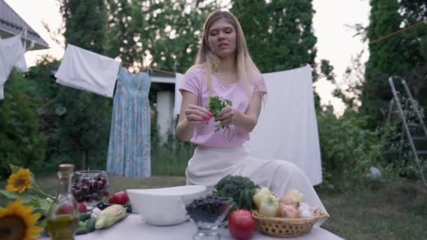 Giovane casalinga fiduciosa che strappa prezzemolo al rallentatore in una ciotola bianca aggiungendo ingredienti per l'insalata. Ritratto di bella donna caucasica magra che cucina cibo salutare seduta sul cortile. — Video Stock