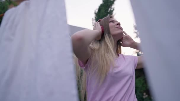 Joven mujer feliz en auriculares bailando a la música con ropa blanca limpia colgando de la cuerda en la parte delantera. Retrato de ama de casa caucásica positiva disfrutando de hobby al aire libre en el patio trasero. — Vídeos de Stock