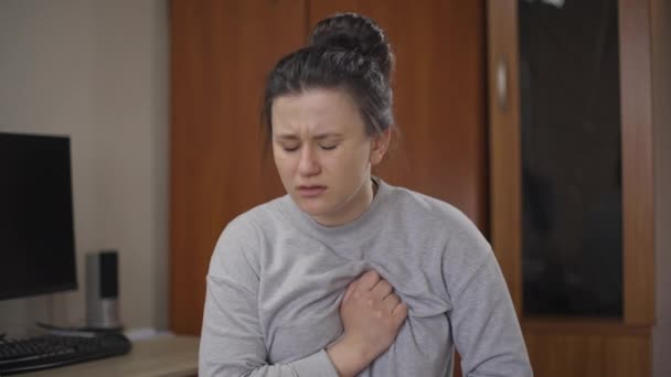 Potret wanita muda dengan nyeri parah di dada duduk di dalam rumah. Wanita sakit Kaukasia sedih dengan serangan jantung menahan napas di dada. Konsep penyakit kardiovaskular. — Stok Video