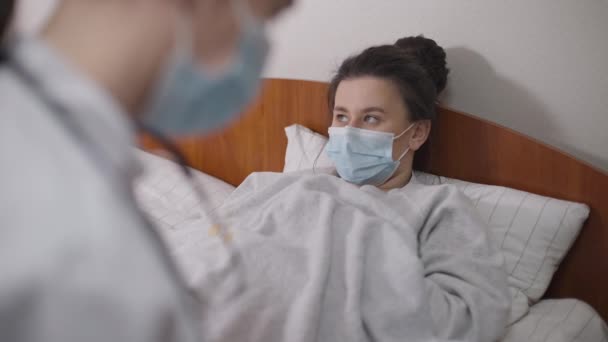 Portret van een jonge vrouw die in bed ligt in het ziekenhuis en praat met een arts die klachten heeft. Ill mooie dame in coronavirus gezichtsmasker in kliniek binnen op Covid-19 pandemie. Geneeskunde en ziekte. — Stockvideo