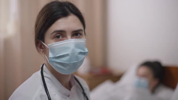 Die traurige Ärztin in Covid-19-Gesichtsmaske in Großaufnahme seufzt, reibt sich die Stirn mit der Hand und blickt in die Kamera. Kopfschuss-Porträt einer überarbeitenden Kaukasierin, die drinnen posiert, verschwommene Patientin im Hintergrund. — Stockvideo