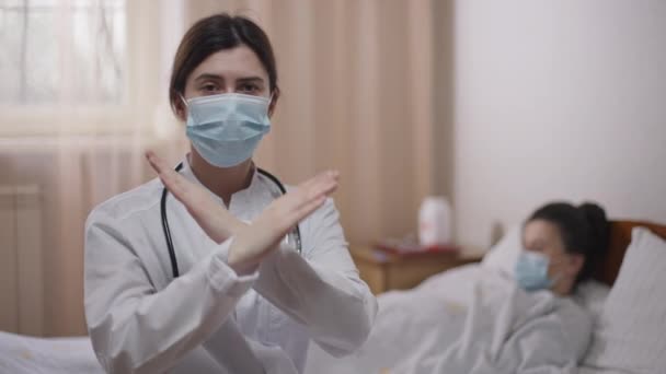 Ernsthaft intelligente junge Ärztin, die die Hände kreuzt, keine Geste, die in die Kamera schaut, mit verschwommenem Patienten, der im Hintergrund auf der Krankenhausstation im Bett liegt. Selbstbewusste Kaukasierin posiert drinnen auf Covid-19. — Stockvideo