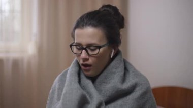 Gözlüklü genç bir kadın yatak odasında oturmuş kulaklıkla müzik dinliyor. Kapalı alanda koronavirüs belirtileri gösteren üzgün Kafkasyalı hasta kadının portresi..
