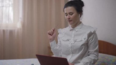 Mutlu, genç bir kadın, kredi kartını kapatıyor, bilgisayarını kapatıyor, gülümsüyor ve yatak odasında oturuyor. İnternetten sipariş veren beyaz bir müşterinin portresi. Çevrimiçi alışveriş kolaylığı.