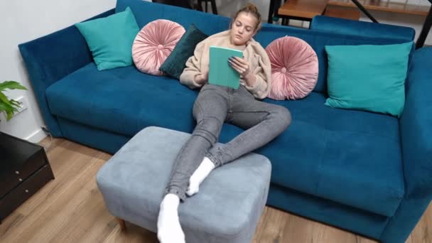 リビングのソファの上に横たわるタブレットの目撃にソーシャルメディアで10代の女の子のメッセージを退屈。週末には屋内でインターネットサーフィンをしている白人青年を撮影した。退屈とライフスタイル. — ストック動画