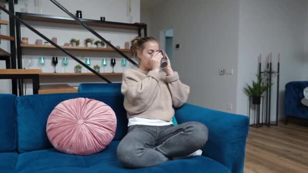 Široký záběr šťastný dospívající dívka pije čaj kávu sedí na gauči doma odpočívá o víkendu. Portrét s úsměvem jistý roztomilý kavkazský teenager těší volný čas uvnitř s teplým nápojem. — Stock video