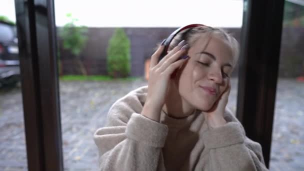 Front view joyful teenage girl singing listening to music in headphones sitting indoors at window smiling. Portrait of relaxed carefree Caucasian teenager enjoying hobby on weekend at home. — Stock Video