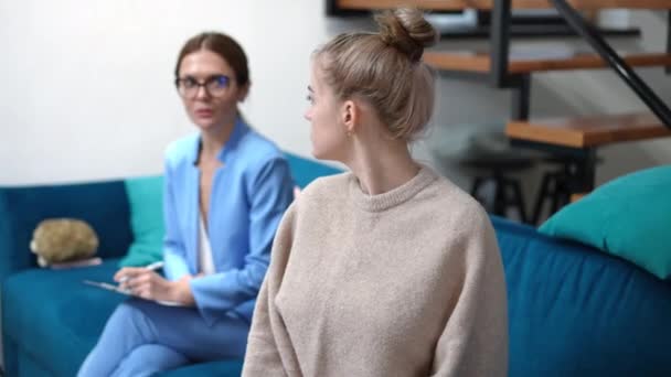 Ontevreden tienermeisje kijkt terug op wazige therapeut die naar camera zucht. Portret van ongelukkige blanke adolescente patiënt die binnen poseert in het psycholoog kantoor. Irriteerbaarheid. — Stockvideo