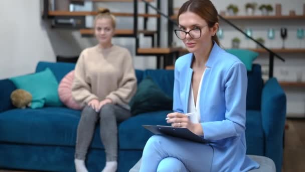 Inteligente caucásica hermosa terapeuta en gafas mirando a la cámara sonriendo con borrosa adolescente cliente de fondo. Retrato de experto psicólogo profesional inteligente posando en la oficina en el interior — Vídeo de stock