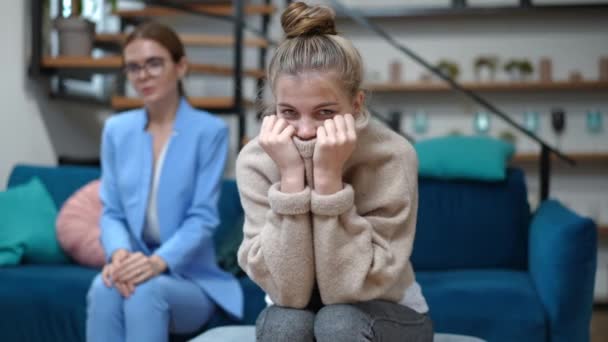 Retrato de adolescente introvertida olhando para câmera cobrindo o rosto inferior com suéter sentado na consulta de psicólogo. Terapeuta caucasiano triste balançando a cabeça no fundo. Conceito de adolescência. — Vídeo de Stock