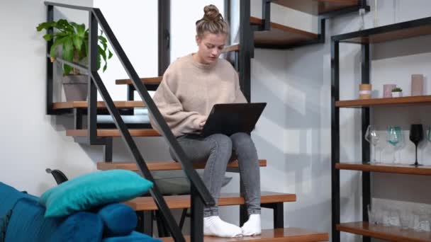 Széles körű portré koncentrált tizenéves lány üzenetküldés online gépelés laptop billentyűzeten ül a lépcsőn beltérben. Elnyelt kaukázusi tinédzser szörfözik otthon az interneten. Modern technológiák. — Stock videók